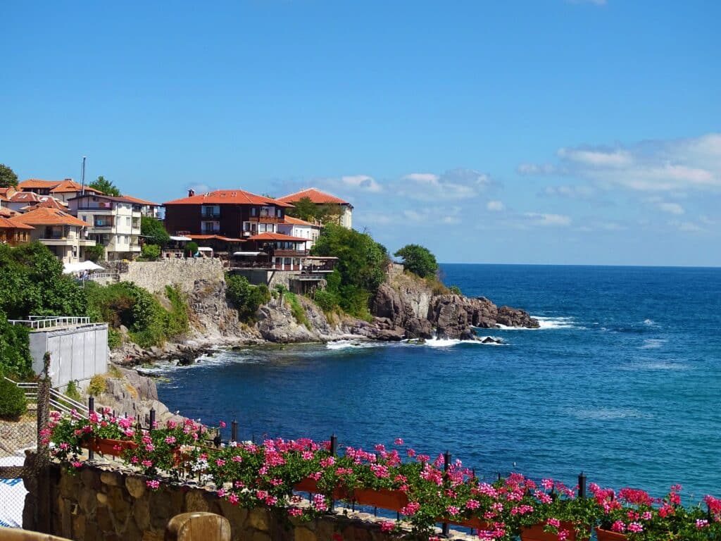 Coastal village with red-roofed houses perched on rocky cliffs overlooking a blue sea; pink flowers line a fence in the foreground under a bright, sunny sky—a perfect escape to explore Bulgaria's Golden Visa: Everything You Need to Know.