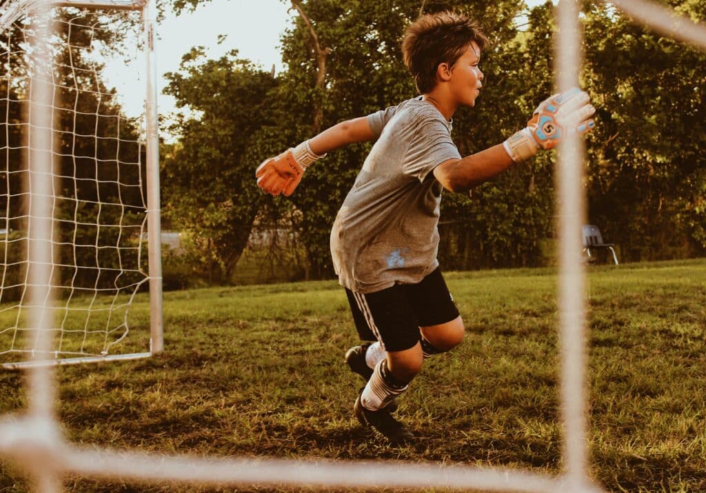 A young football goalkeeper wearing gloves and a grey shirt runs in front of the goal on a grassy pitch, focused on the game—a great example of the benefits of sport in schools—with trees and sunlight in the background.