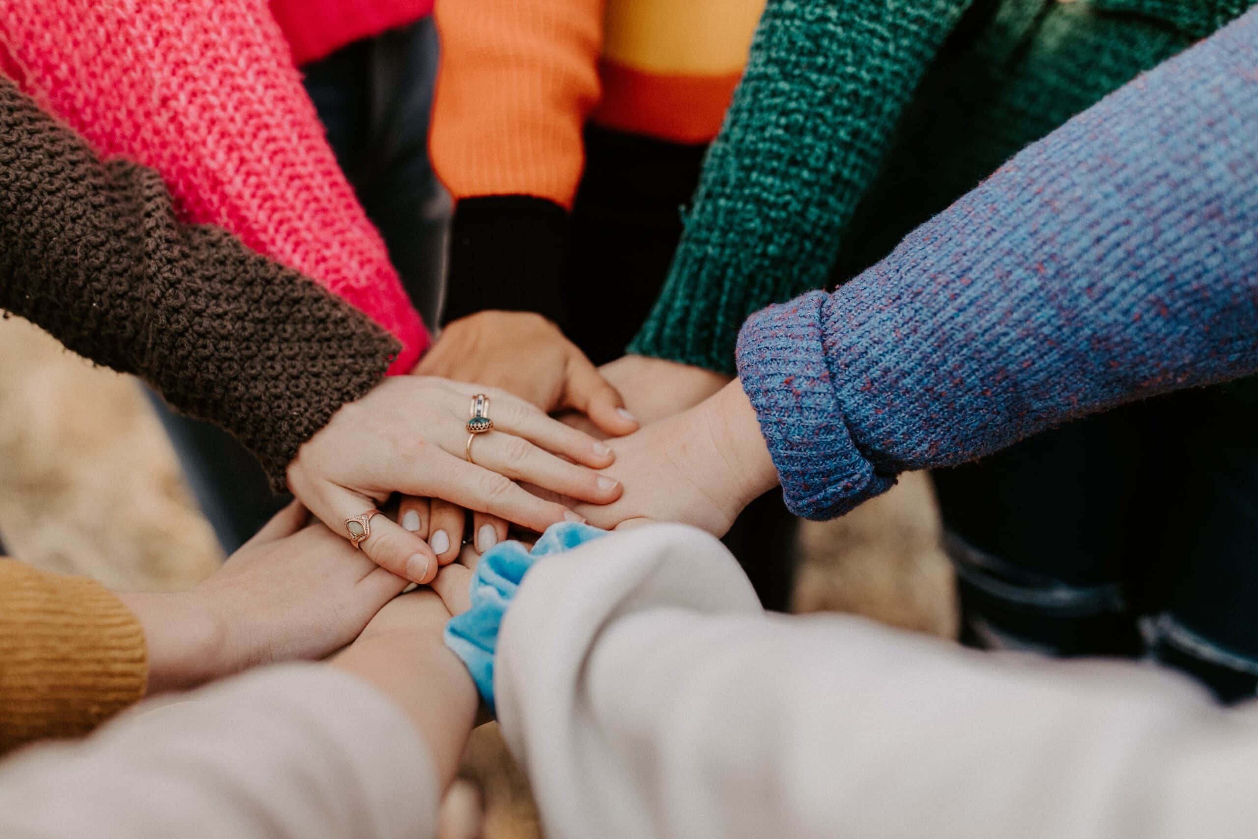 Several people wearing colourful jumpers join hands in the centre of a circle, symbolising unity, teamwork, and support. This moment highlights why community involvement is important for children—helping them build connections and a sense of belonging.