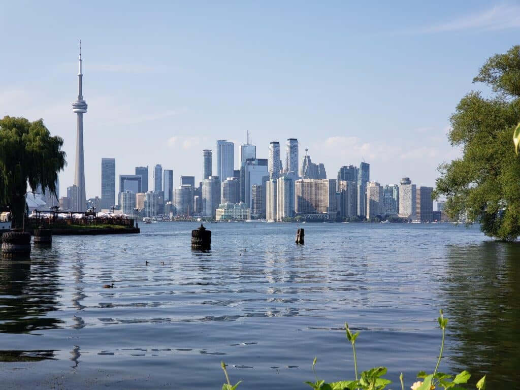 Toronto skyline viewed across the water on a sunny day, featuring the CN Tower and skyscrapers where many choose to live and work, with trees framing the foreground and gentle ripples glistening on the lake.
