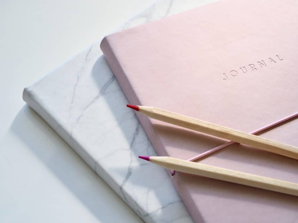 A pink diary with “JOURNAL” embossed on the cover and a white marble-patterned notebook underneath, with two coloured pencils resting diagonally on top—a perfect setup to inspire why you should encourage your child to start journalling.