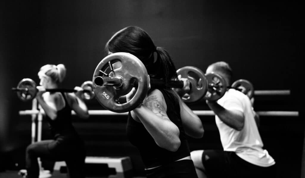 Three people in workout attire perform squats with barbells in a black and white gym scene. The focus is on the centre athlete, showcasing how to level up your workout style with gym clothes that will boost your confidence.