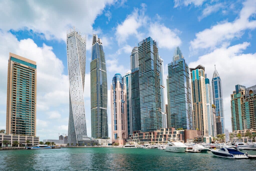 Modern skyscrapers and twisted buildings line the waterfront of a marina, creating a vibrant place to live and work, with several yachts and boats moored in turquoise water under a blue sky with scattered clouds.