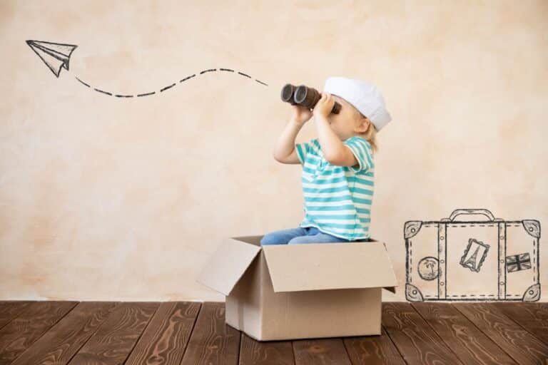 A child in a striped shirt and sailor hat sits in a cardboard box, looking through binoculars. A drawn paper aeroplane and suitcase decorate the background, highlighting imagination, adventure, and independent play.