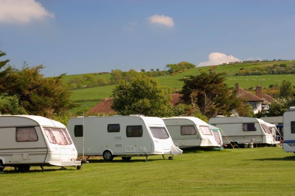 Several white caravans are parked on a grassy field among rolling hills and scattered trees, making it an ideal spot to learn how to stay safe in your caravan under a blue sky with light clouds.