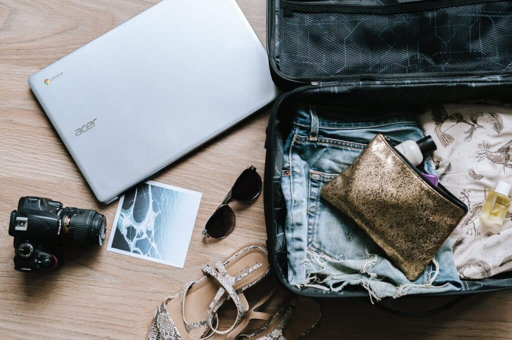 Open suitcase on the floor with denim shorts, a gold pouch, sandals, sunglasses, travel-size toiletries, a camera, a laptop, and a printed photo arranged beside it, capturing the excitement of preparations for an upcoming journey.
