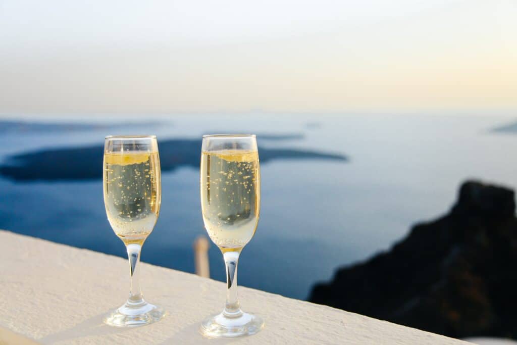 Two champagne glasses with fizzy drinks rest on a white ledge, capturing the perfect mini-moon moment as they overlook a scenic view of the sea and distant islands at sunset.