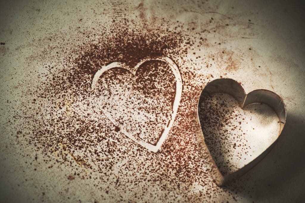 A heart shaped cookie cutter next to some cocoa powder with a heart shaped mark in the powder
