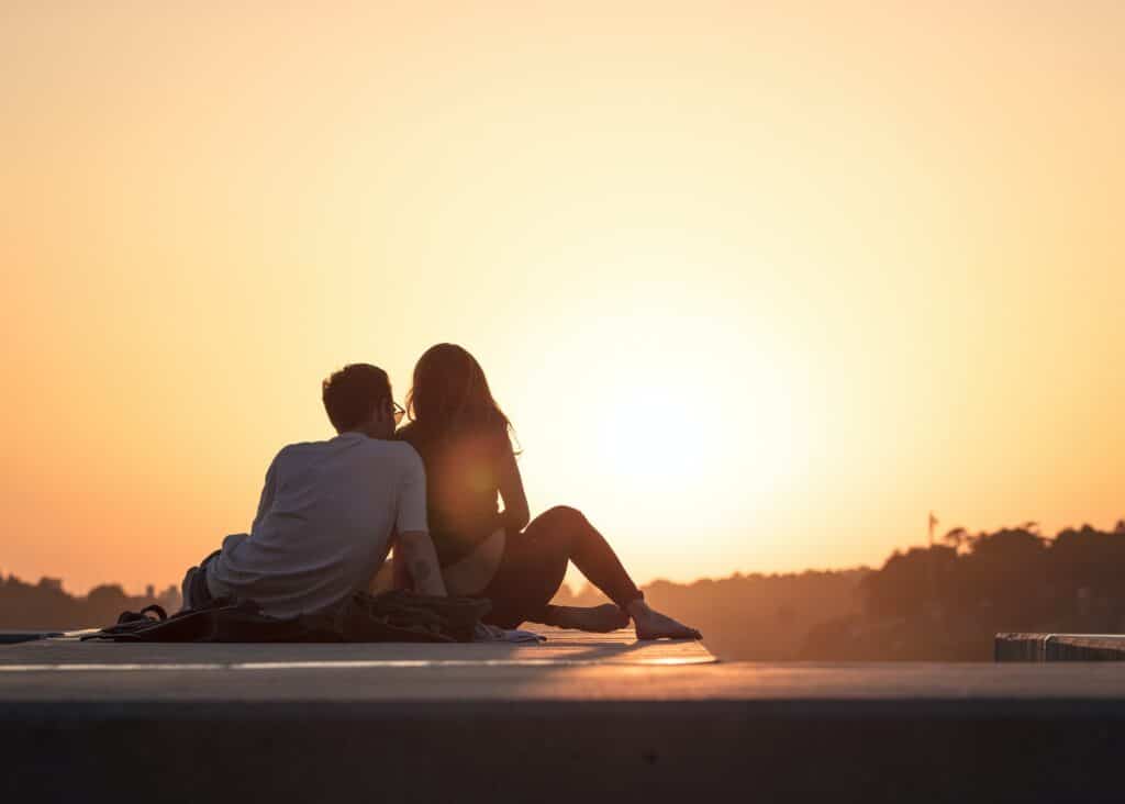 A couple sit close together on a ledge, watching the sunset during their mini-moon. The sky is a warm orange, and their silhouettes are outlined against the bright sun on the horizon.