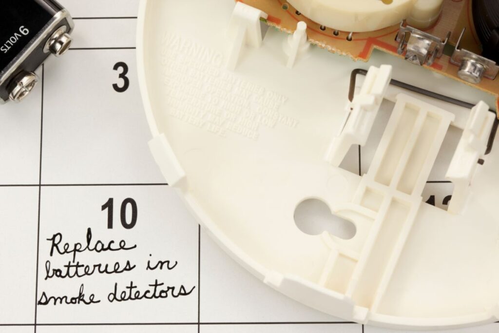 An open smoke detector sat on top of a diary next to a new battery. On the 10th of the month it says to replace battery in the smoke detector