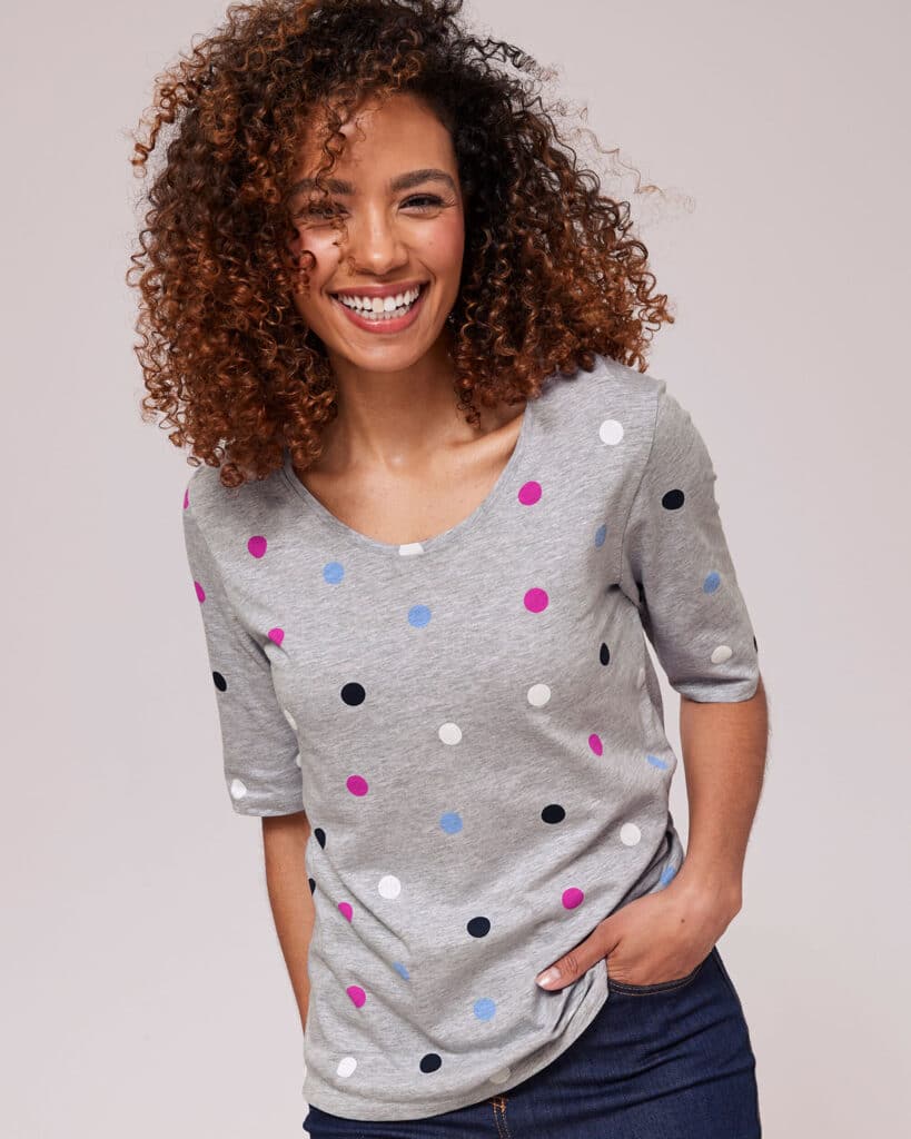 A woman with curly hair smiles in comfortable clothing—a grey shirt with colourful polka dots in pink, blue, black, and white. With one hand in her pocket, she stands relaxed against a plain light background.