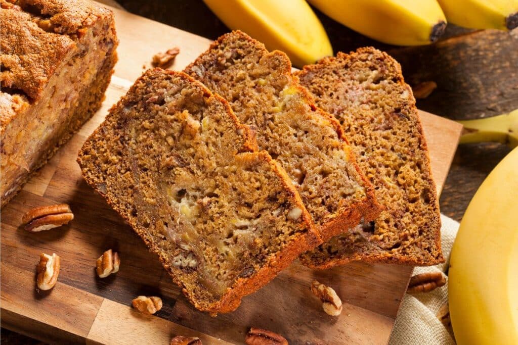 Two slices of banana bread with visible nuts rest on a wooden chopping board, surrounded by chopped pecans and whole bananas—the perfect treat for baking with plant milk.