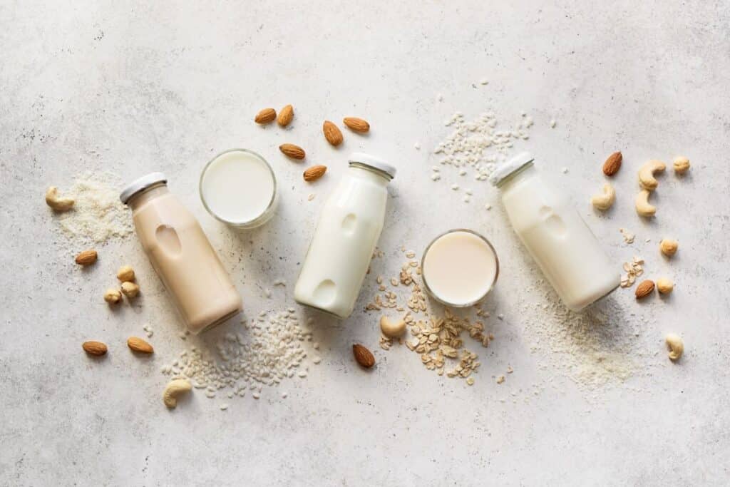 3 bottles of different types of plant milk laid on their sides surrounded by nuts and two glasses of milk