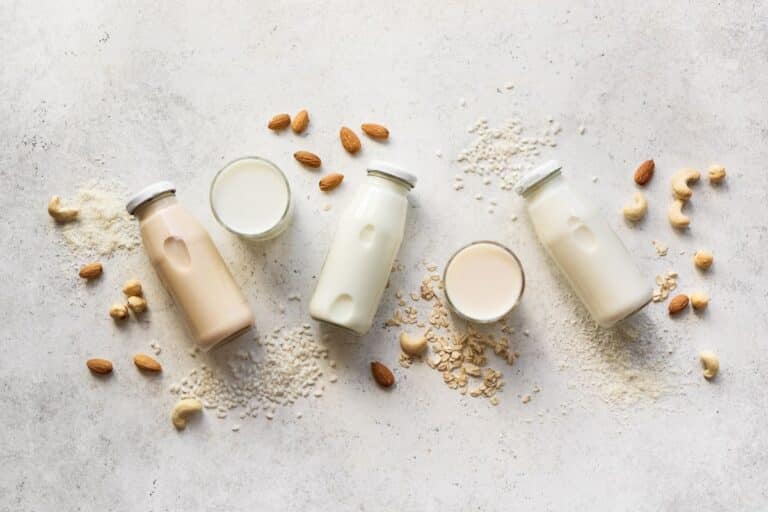 3 bottles of different types of plant milk laid on their sides surrounded by nuts and two glasses of milk