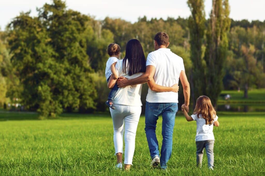 A foster carer in Yorkshire walks with two young children and another adult across a green field towards trees and a pond, enjoying a sunny day outdoors.