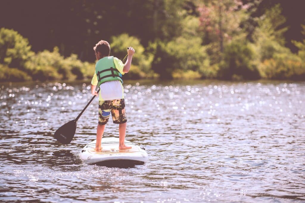 A child wearing a life jacket and colourful shorts stands on a paddleboard, showing how to get the most out of what remains of summer, holding a paddle on a calm, sunlit lake surrounded by greenery.