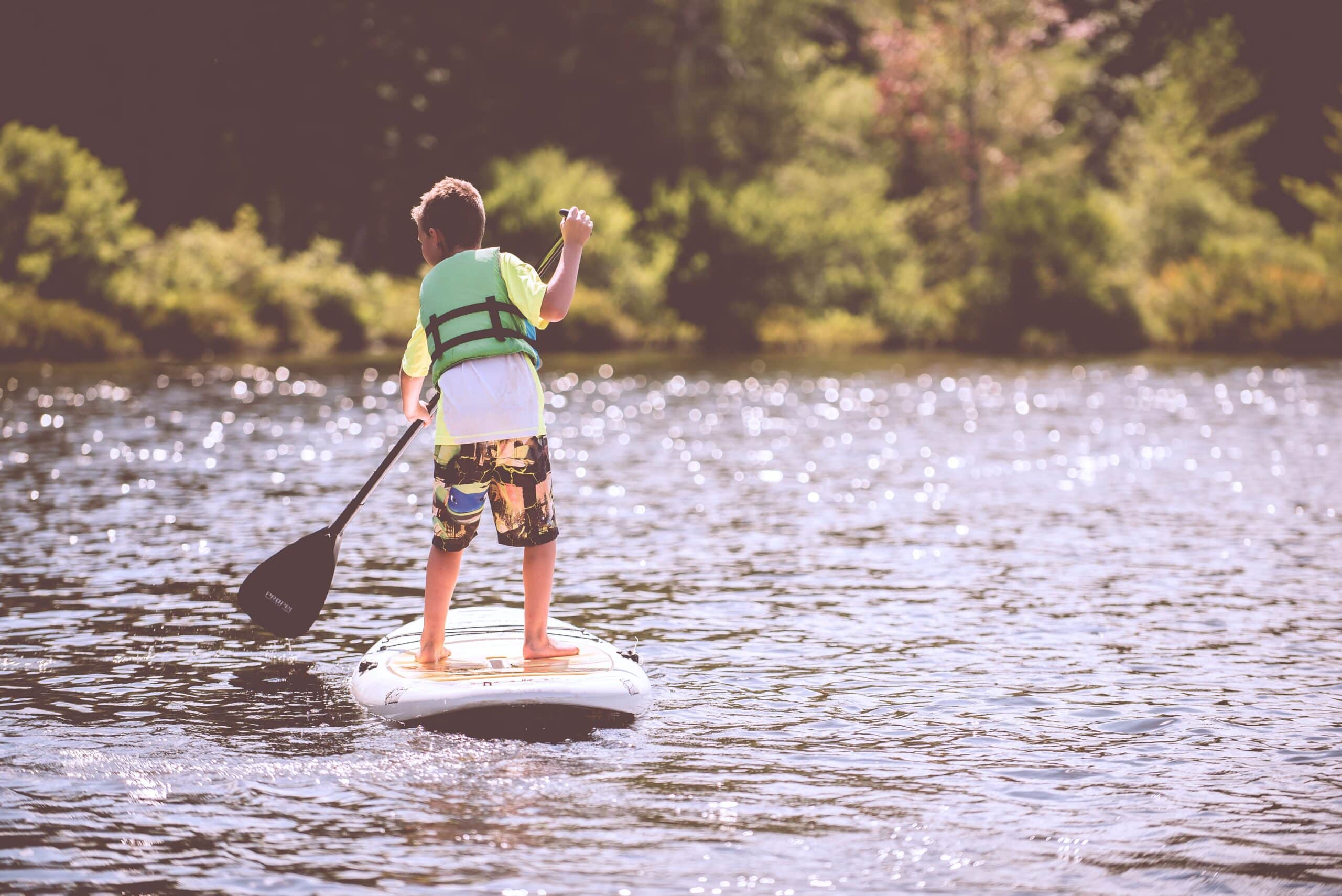 A child wearing a life jacket and colourful shorts stands on a paddleboard, showing how to get the most out of what remains of summer, holding a paddle on a calm, sunlit lake surrounded by greenery.