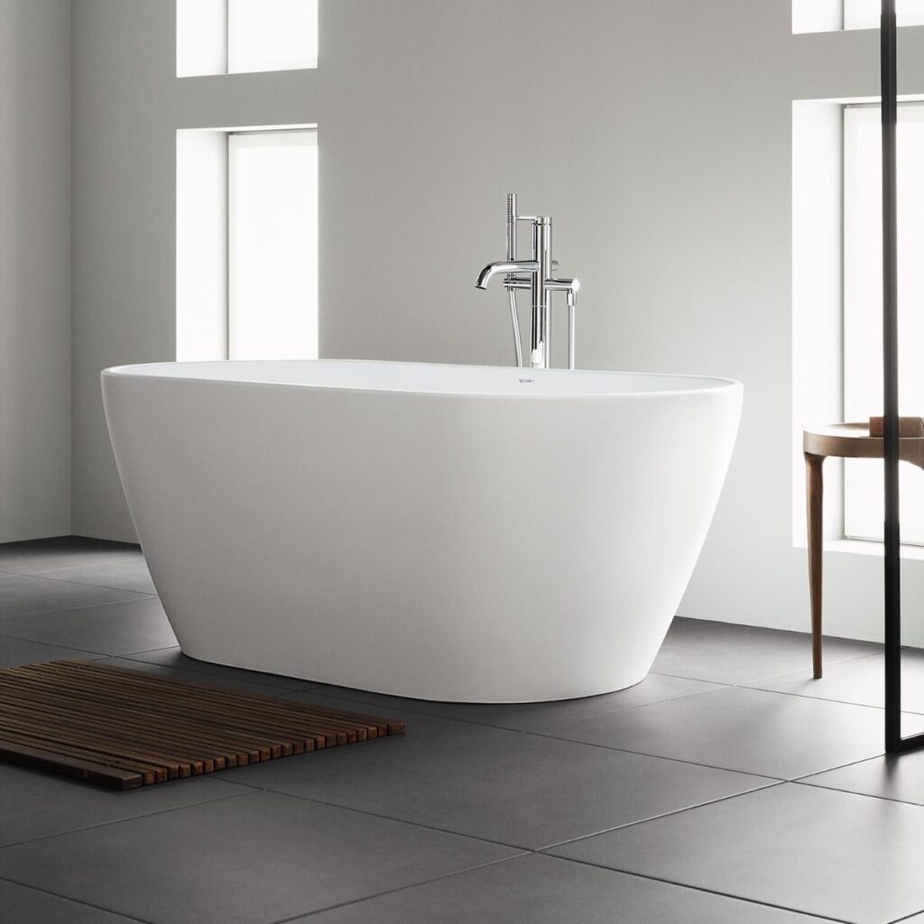 Modern white freestanding bath with chrome tap in a minimalist bathroom, featuring large windows, grey tiled floor, a wooden bath mat, and a small wooden table. Natural light brightens the bathroom space.