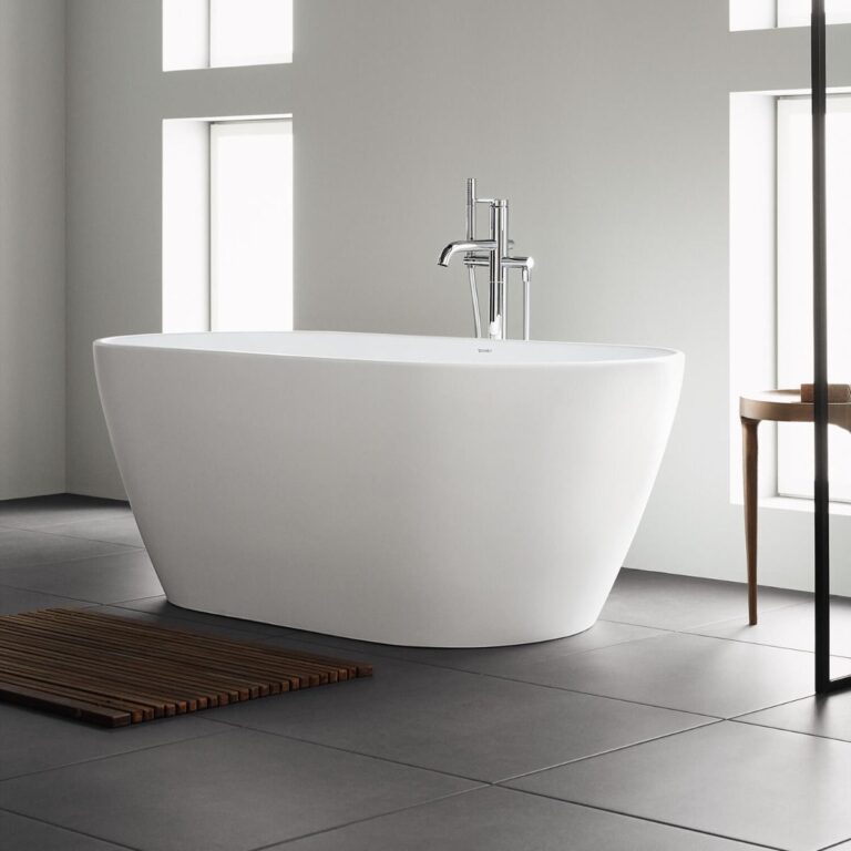 Modern white freestanding bath with chrome tap in a minimalist bathroom, featuring large windows, grey tiled floor, a wooden bath mat, and a small wooden table. Natural light brightens the bathroom space.