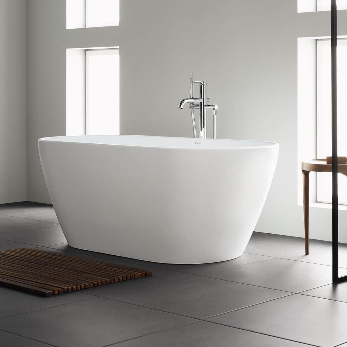 Modern white freestanding bath with chrome tap in a minimalist bathroom, featuring large windows, grey tiled floor, a wooden bath mat, and a small wooden table. Natural light brightens the bathroom space.