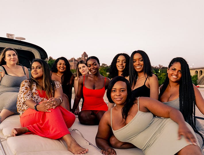 A group of nine women sit and relax together on a boat at sunset, smiling in stylish shaping dresses and enjoying each other’s company. They bask in the warm glow, with trees and buildings visible in the background.