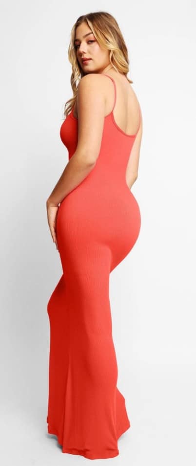 Woman standing in a side pose, wearing a long, sleeveless, figure-hugging red shaper dress with thin straps. She has long, wavy blonde hair and a neutral expression against a plain white background.