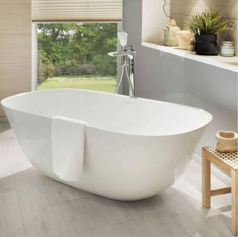 A modern white freestanding bath with a towel draped over the side sits in a bright bathroom featuring light wood floors, lush plants, and a large window overlooking a green garden.