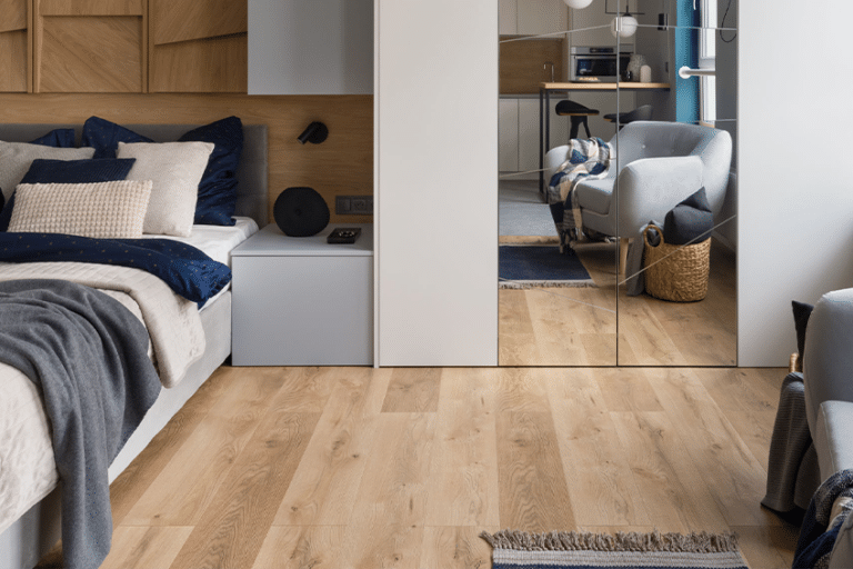 A modern bedroom with vinyl flooring, a bed with blue and beige bedding, a grey bedside table, large mirrored wardrobe, grey armchair, and a woven basket. The room is tidy and well-lit with natural light.