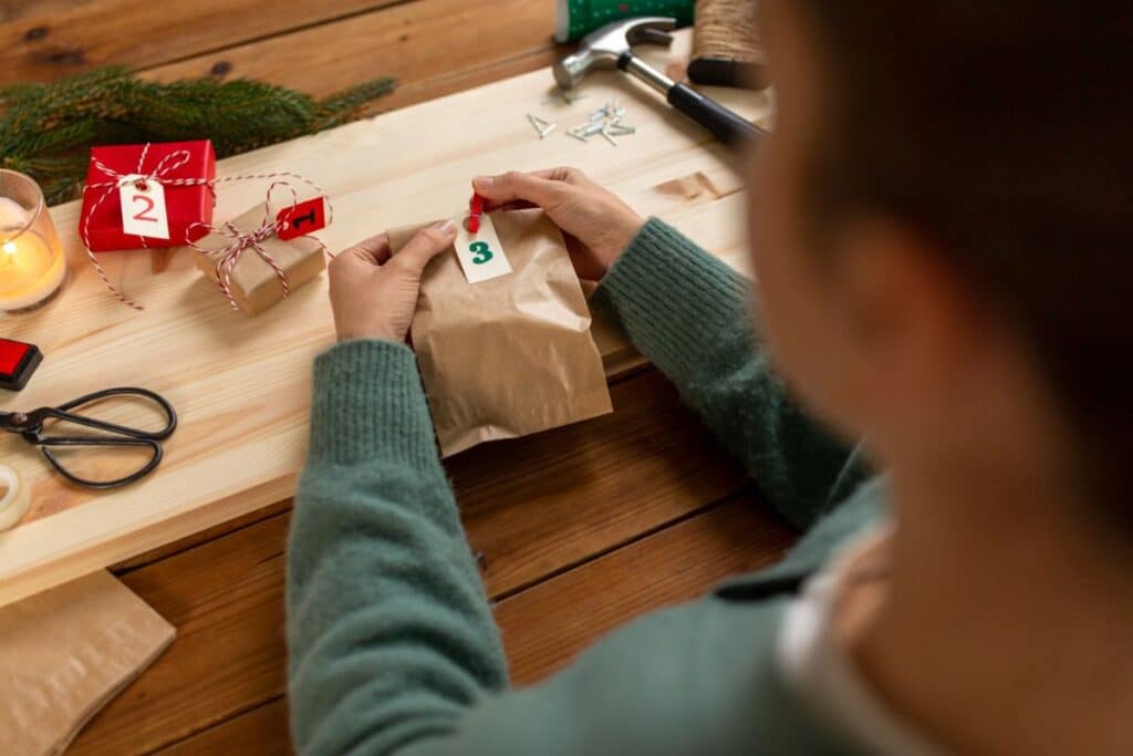 A person wearing a green jumper holds a brown paper bag labelled "3" from an Advent Calendar on a wooden table, surrounded by wrapped presents, scissors, string, and a candle.