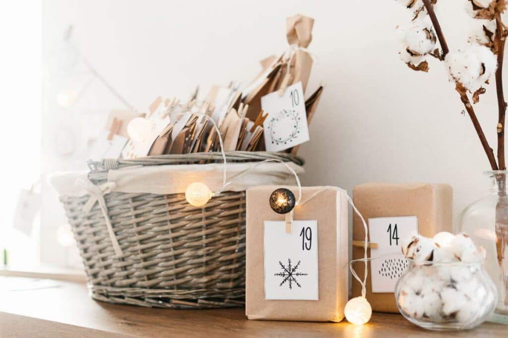 A wicker basket filled with brown paper-wrapped Advent Calendar presents sits on a wooden surface. Two numbered presents, "14" and "19," are in front, decorated with fairy lights and dried cotton stems.