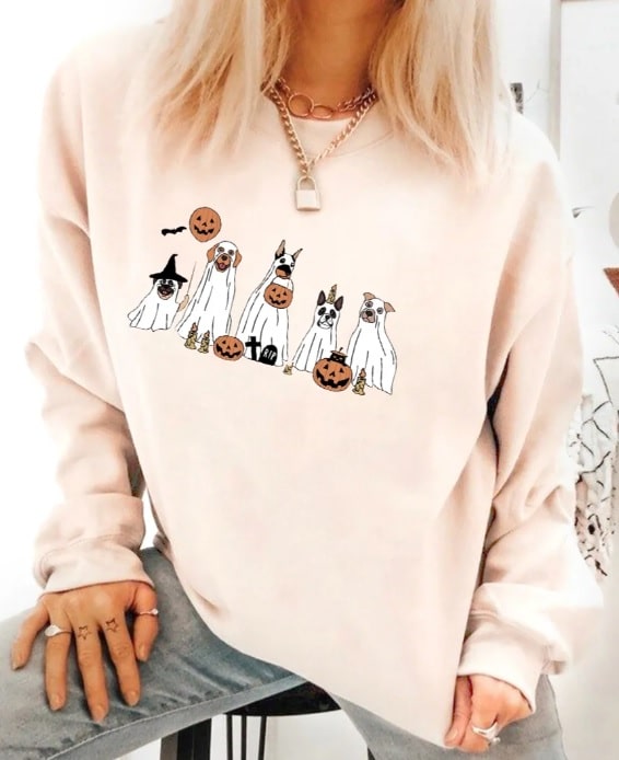 A person wearing an oversized, light-coloured sweatshirt featuring five dogs dressed as ghosts holding pumpkins and Halloween props. The person’s face is not shown.