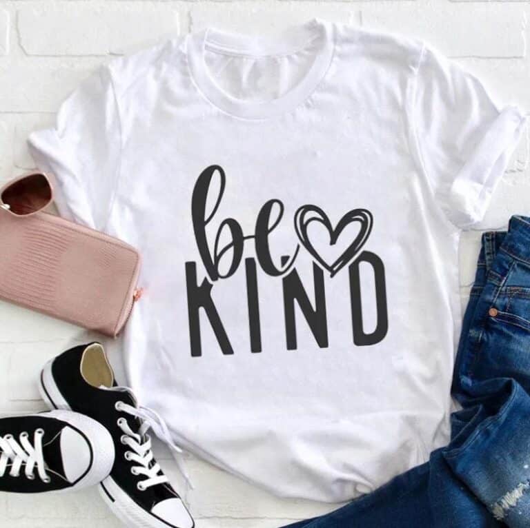 A white teacher T-shirt with the phrase “be KIND” in bold black letters and a heart-shaped "e" is laid out next to blue jeans, black-and-white trainers, and a pale pink zippered pouch on a white surface.
