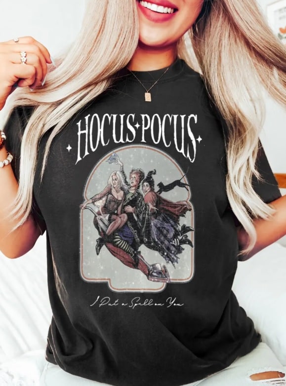 A woman with long blonde hair smiles whilst wearing an oversized "Hocus Pocus" T-shirt featuring illustrated witches riding a broomstick and the text "I Put a Spell on You.