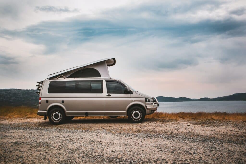 A silver camper van with its pop-up roof extended is parked on a gravel area by a lake, with hills in the background under a cloudy sky—perfect for spotting the signs it's time to change your vehicle and upgrade your adventure.