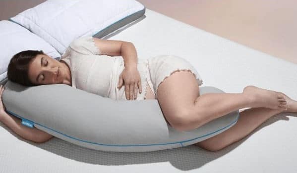 A pregnant woman lies on her side in bed, hugging a large, grey, U-shaped body pillow, with her eyes closed and appearing comfortable and relaxed.