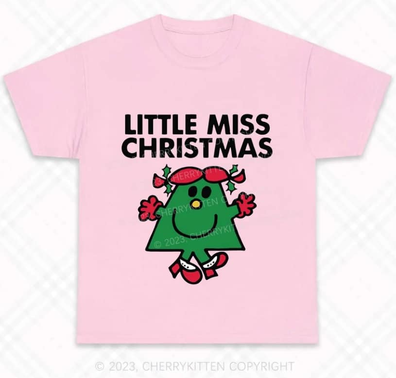 A light pink T-shirt in a festive style featuring the text "LITTLE MISS CHRISTMAS" above a cartoon green Christmas tree character with red mittens, boots, and baubles as earrings.