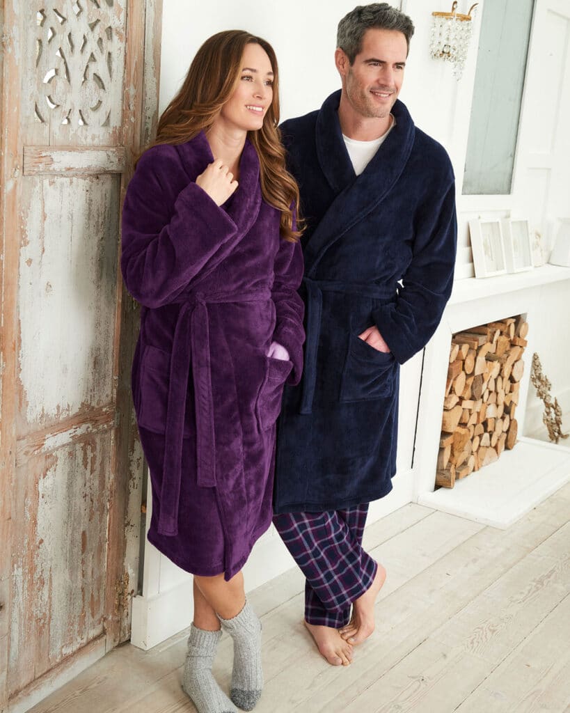 A smiling woman in a purple dressing gown and grey socks stands beside a smiling man in a navy dressing gown and checked pyjama trousers, both showcasing cosy knitwear indoors near a white fireplace and stacked firewood.