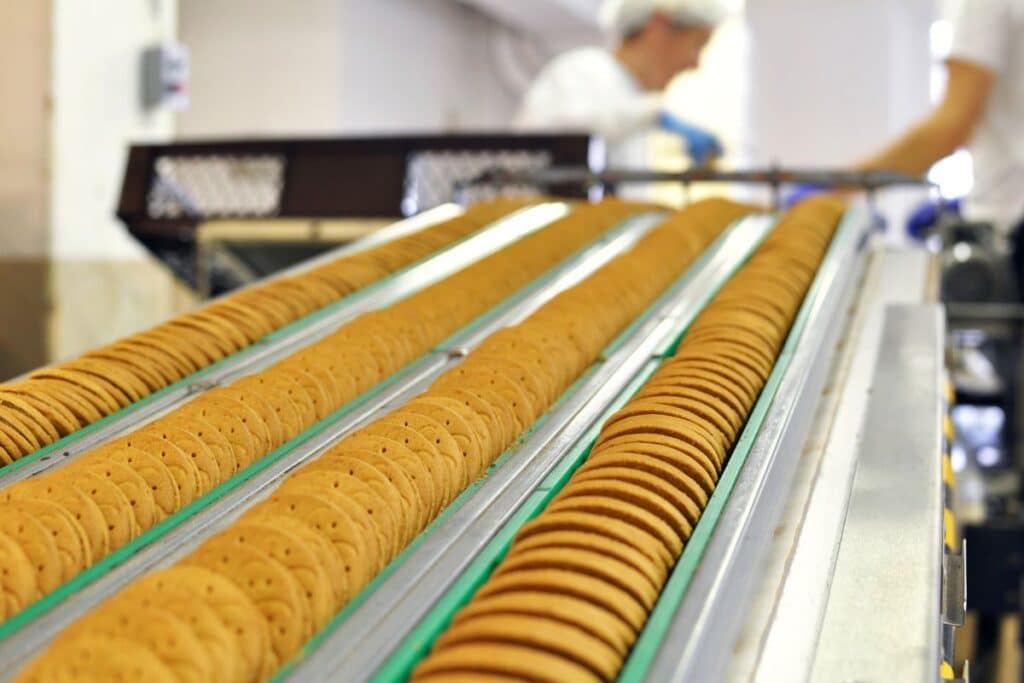 Rows of round biscuits move along a conveyor belt in a food factory, where workers in white uniforms and hairnets oversee the food preparation process in the background.
