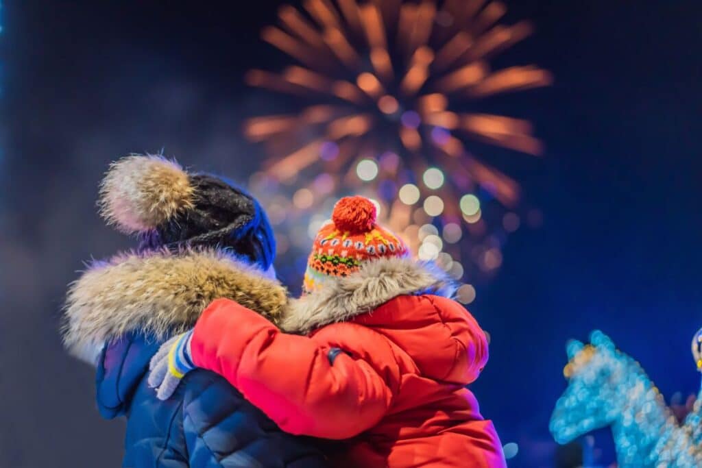 Two people bundled in winter coats and colourful hats embrace while watching a vibrant fireworks display, enjoying one of many outdoor winter activities. Bright lights and a carousel horse add to the festive atmosphere in the background.