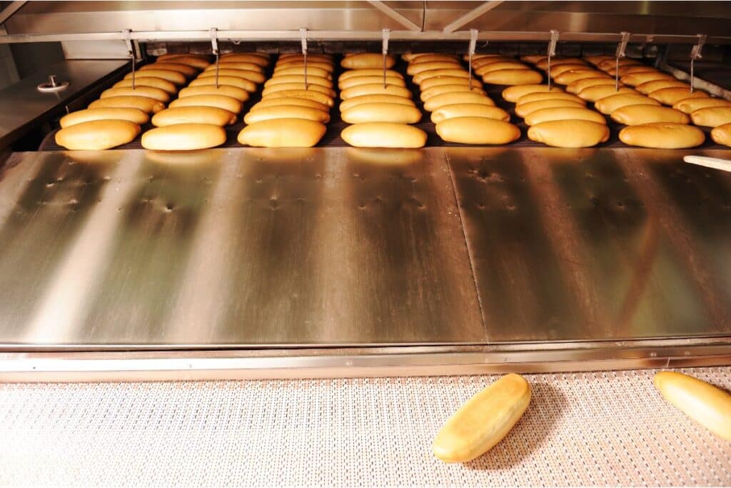Rows of fresh loaves of bread move along a bakery conveyor belt during food preparation, with one loaf separated from the group at the front edge of the belt.