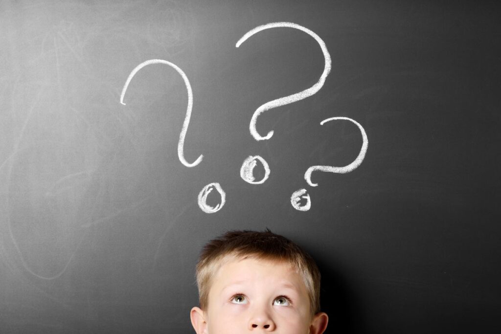 A young boy looks up thoughtfully in front of a blackboard with three large question marks above his head, highlighting the learning challenges and curiosity he faces.
