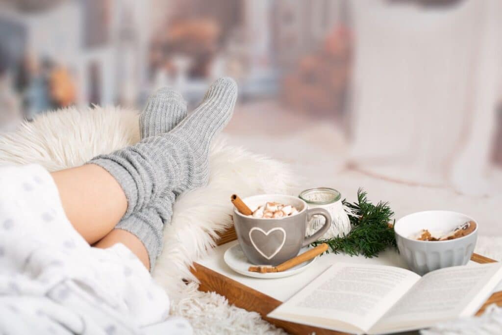 A person wearing cosy grey knitwear socks relaxes on a fluffy blanket with hot chocolate topped with marshmallows, an open book, and a small bowl of snacks on a wooden tray, creating a warm and cosy atmosphere.