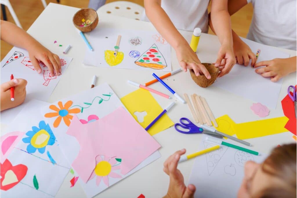 Children’s hands working on colourful crafts at a table, with drawings, coloured paper, scissors, glue, lollipop sticks, and felt-tip pens spread out, sparking creativity as they create various art projects.
