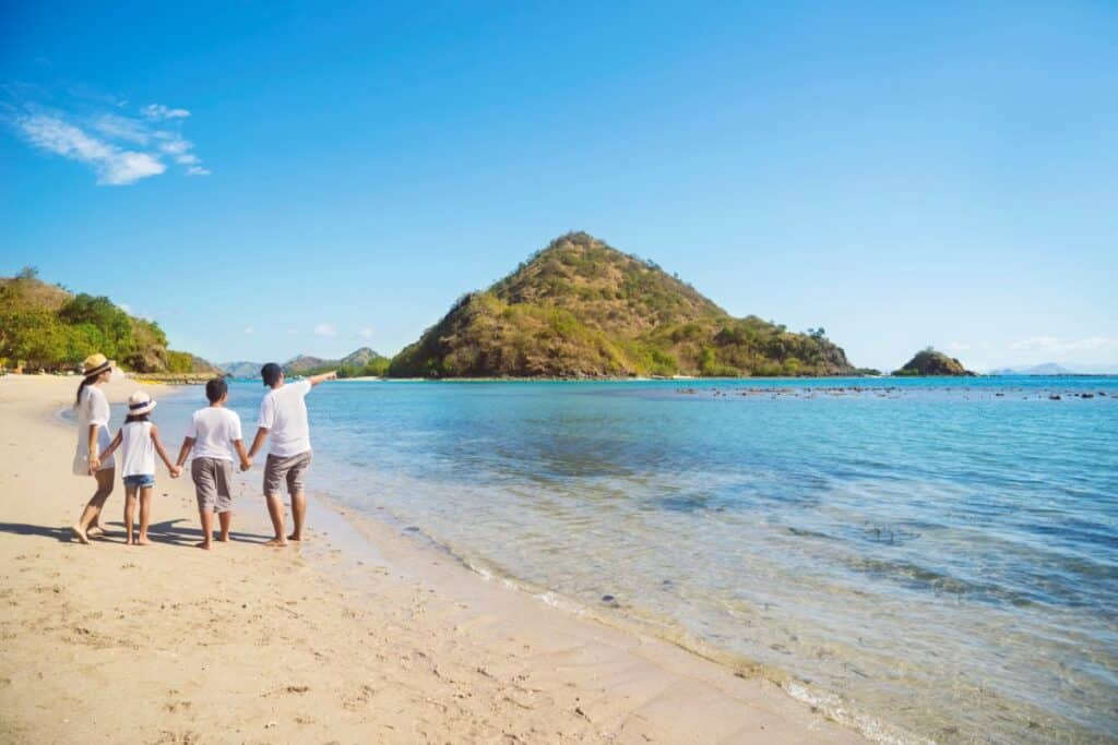 A family of four walks along a sandy beach, holding hands and looking towards a green hill across clear blue water—enjoying the once-in-a-lifetime family holiday they spent years saving for.