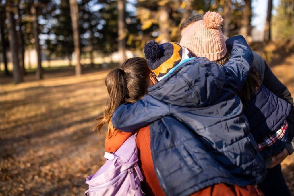 Three children wearing winter jackets and hats share a group hug outdoors in a park with autumn leaves, capturing the warmth and togetherness often found in fostering in Yorkshire. Trees stand tall in the background.
