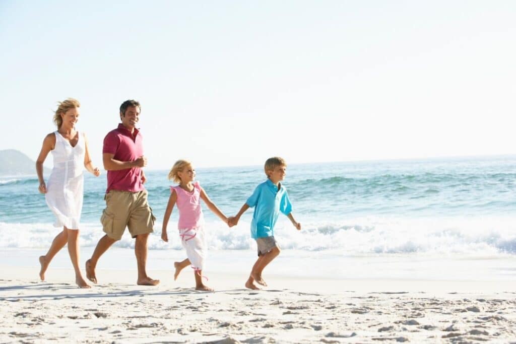 A family of four—two adults and two children—runs joyfully along the shoreline of a sunny beach, enjoying their family holiday with ocean waves and blue sky in the background.