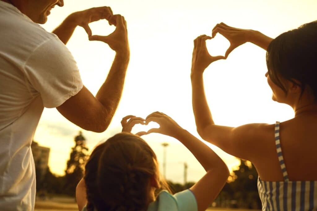 A man, woman, and child stand outdoors at sunset on a family holiday, facing the sun and making heart shapes with their hands above their heads. The scene captures a warm, loving moment in silhouette.