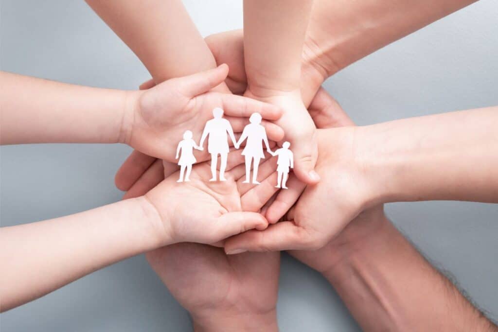 Several hands of adults and children are stacked together, holding a white paper cut-out of a family with two adults and two children, symbolising unity, support, family care, and the importance of foster care.