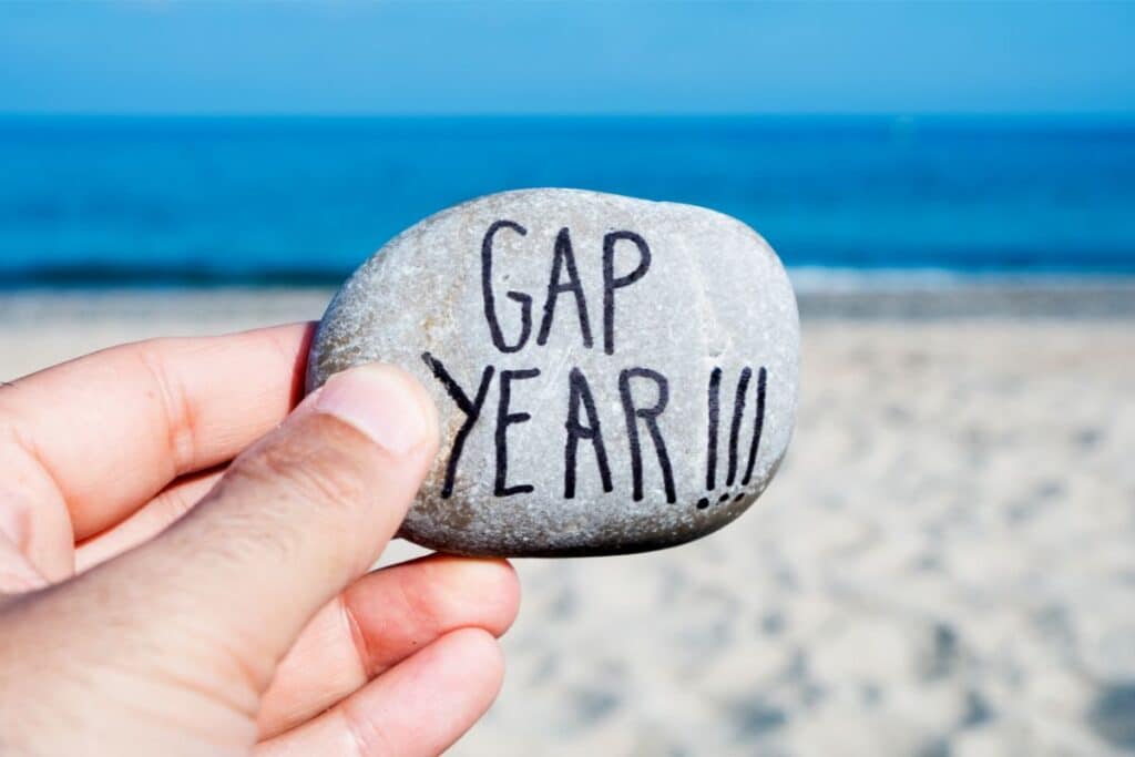 A hand holds a smooth stone with "GAP YEAR!!!" written on it, capturing the spirit of a gap year adventure against the backdrop of a sandy beach and blue sea.
