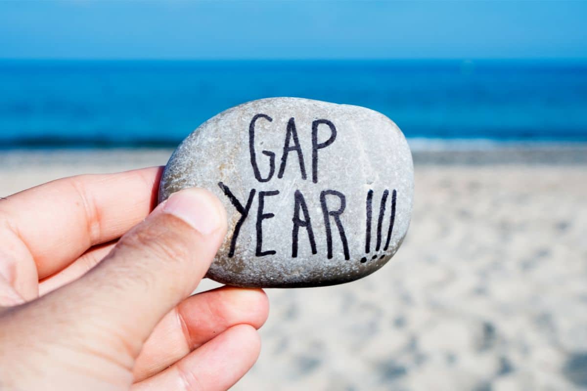 A hand holds a smooth stone with "GAP YEAR!!!" written on it, capturing the spirit of a gap year adventure against the backdrop of a sandy beach and blue sea.